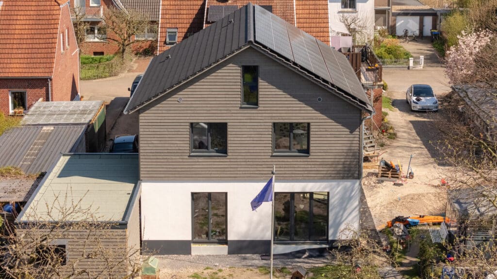 Ein modernes, zweistöckiges Fertighaus aus Holz mit dunklem Dach, Solarpanelen und großen Fenstern steht für nachhaltiges Wohlfühlwohnen. Vor dem Haus zeigt ein Flaggenmast Individualität; umgeben von weiteren Häusern und geparkten Autos.