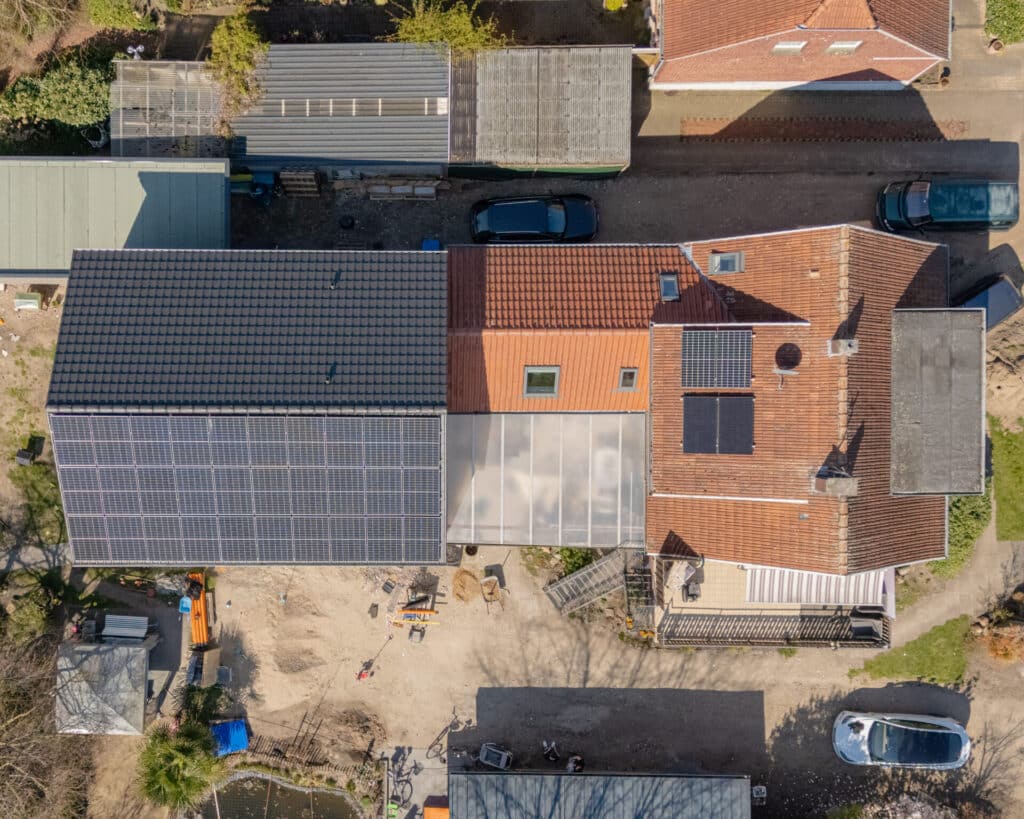 Luftaufnahme von zwei verbundenen Holzhäusern aus einem nachhaltigen Bauprojekt, deren Dächer nahezu vollständig mit Solarpanels bedeckt sind. Umgeben von Autos, Nebengebäuden und einem liebevoll angelegten Garten mit Teich strahlt das Ensemble Behaglichkeit aus.