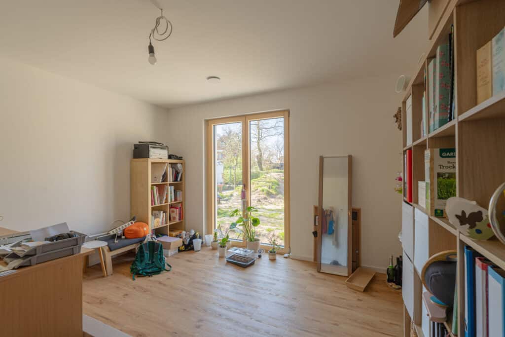 Ein heller, einladender Raum in einem Fertighaus aus Holz überzeugt mit warmen Holzböden, maßgefertigten Bücherregalen und liebevoll platzierten Pflanzen. Großzügige Fenster schenken natürliches Licht und eröffnen den Blick ins Grüne - für ein behagliches Zuhause.