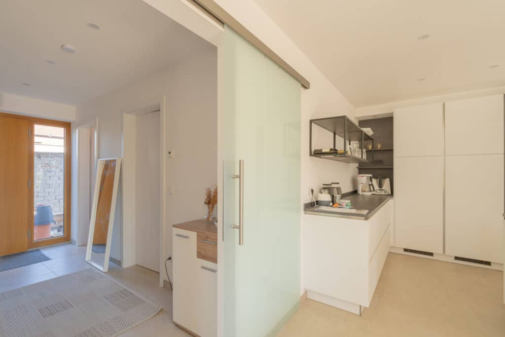 Diese moderne Küche in einem Fertighaus aus Holz vereint weiße Schränke, hochwertige Edelstahlgeräte und eine sanft gleitende Glastür. Durch den hölzernen Eingangsbereich fällt viel Tageslicht und sorgt für eine warme, behagliche Atmosphäre.