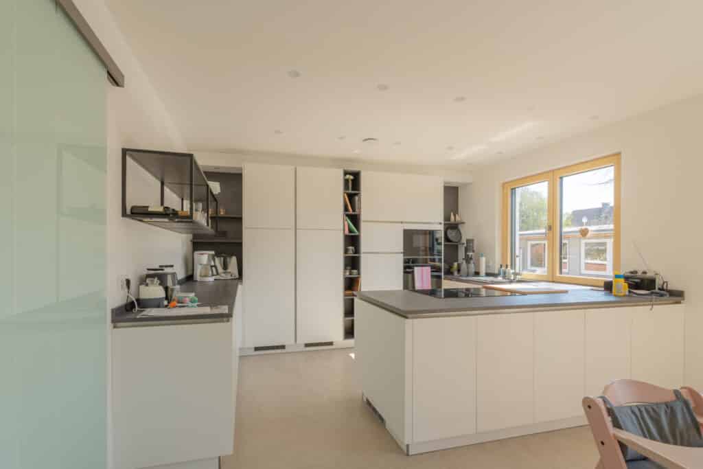 Moderne Küche mit weißen Schränken, großzügiger Kochinsel und grauer Arbeitsplatte - perfekt für Wohlfühlatmosphäre im Holz-Fertighaus. Offene Regale und hochwertige Einbaugeräte schaffen Komfort; ein Hauch Rosa durch einen Stuhl im Vordergrund.