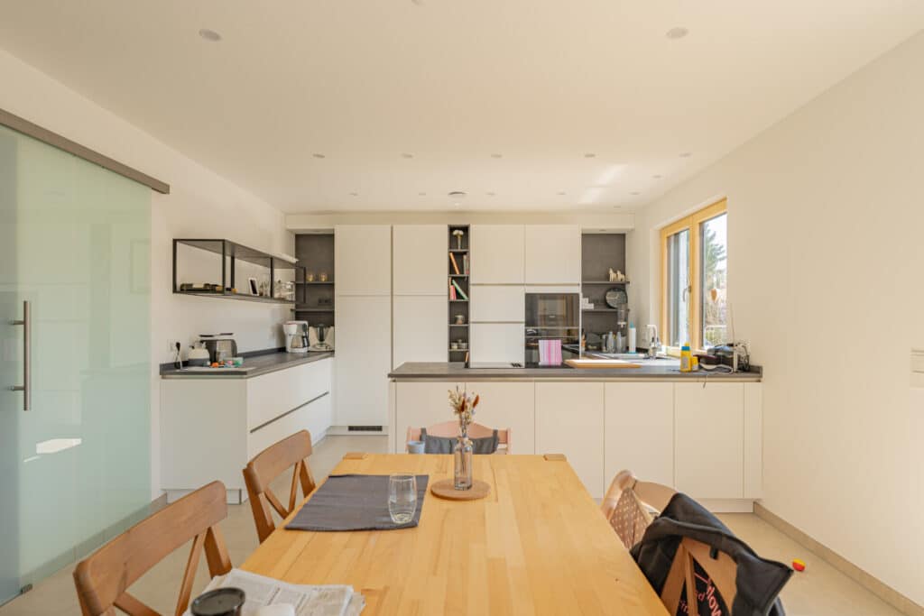 Helle, moderne Küche mit weißen Schränken, integrierten Geräten und einem Holztisch für vier Personen - ein inspirierendes Beispiel für ein behagliches Fertighaus aus Holz. Sonnenlicht betont die hochwertigen Details und das wohnliche Ambiente.
