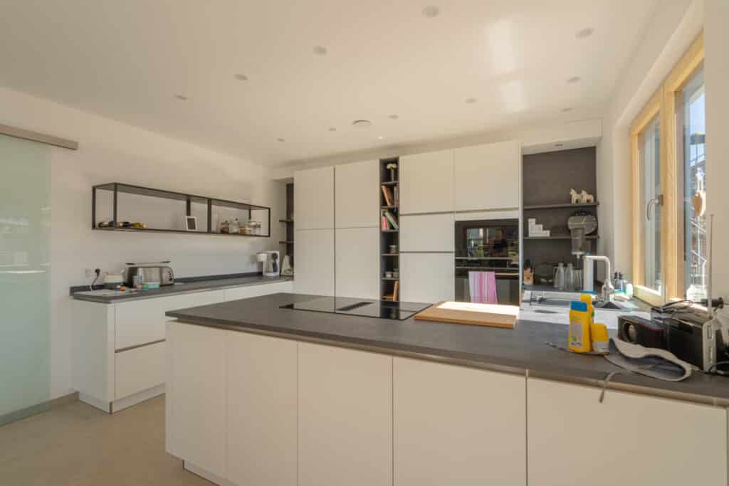 Zeitgemäße Küche mit weißen Schränken, grauen Arbeitsflächen und offenem Regal - einladend und lichtdurchflutet durch ein großes Fenster. Natürliche Materialien und klares Design schaffen Behaglichkeit im nachhaltigen Fertighaus aus Holz.