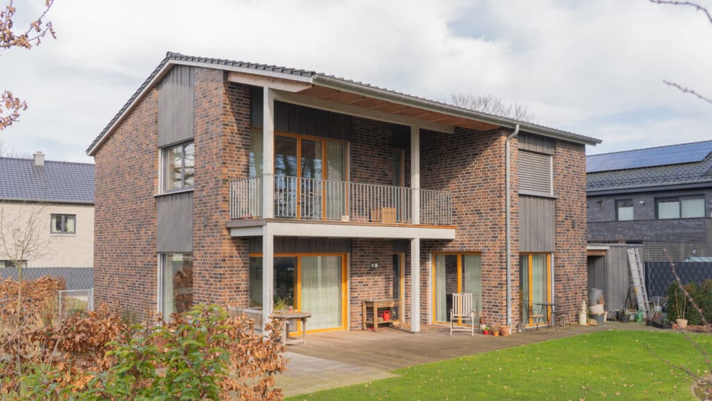 Ein modernes, zweistöckiges Holzhaus kombiniert nachhaltige Bauweise mit stilvollen Ziegelwänden, großen Fenstern und warmen Holzrahmen. Die überdachte Balkonterrasse bietet Blick auf einen grünen Garten und strahlt Behaglichkeit aus.