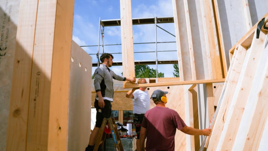 Drei Männer arbeiten gemeinsam an der Montage eines modernen Fertighauses aus Holz unter blauem Himmel. Im Hintergrund stehen Bäume und Baugerüste - nachhaltige Materialien und zeitgemäßes Design schaffen hier wohnlichen Lebensraum.