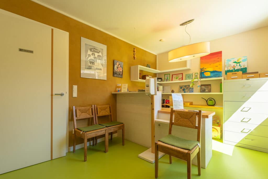 Ein heller, einladender Warteraum mit grünem Boden, Holzstühlen mit grünen Polstern und liebevoller Wandkunst. Die Empfangstheke beherbergt stilvolle Deko und Akten - ein inspirierendes Beispiel für behagliches Wohnen im modernen Holz-Fertighaus.