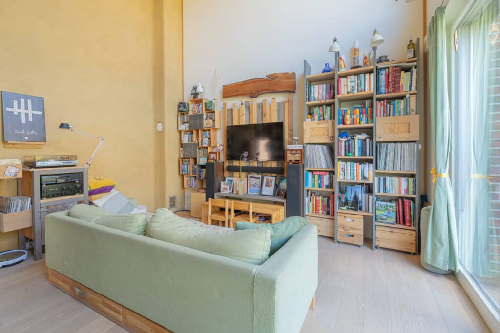 Ein einladendes Wohnzimmer mit hellgrünem Sofa, das sich zur TV-Wand richtet, umgeben von Holzregalen voller Bücher und Deko - ein Sinnbild für wohnliches Wohlfühlambiente. Perfekte Inspiration für nachhaltige Fertighäuser aus Holz.