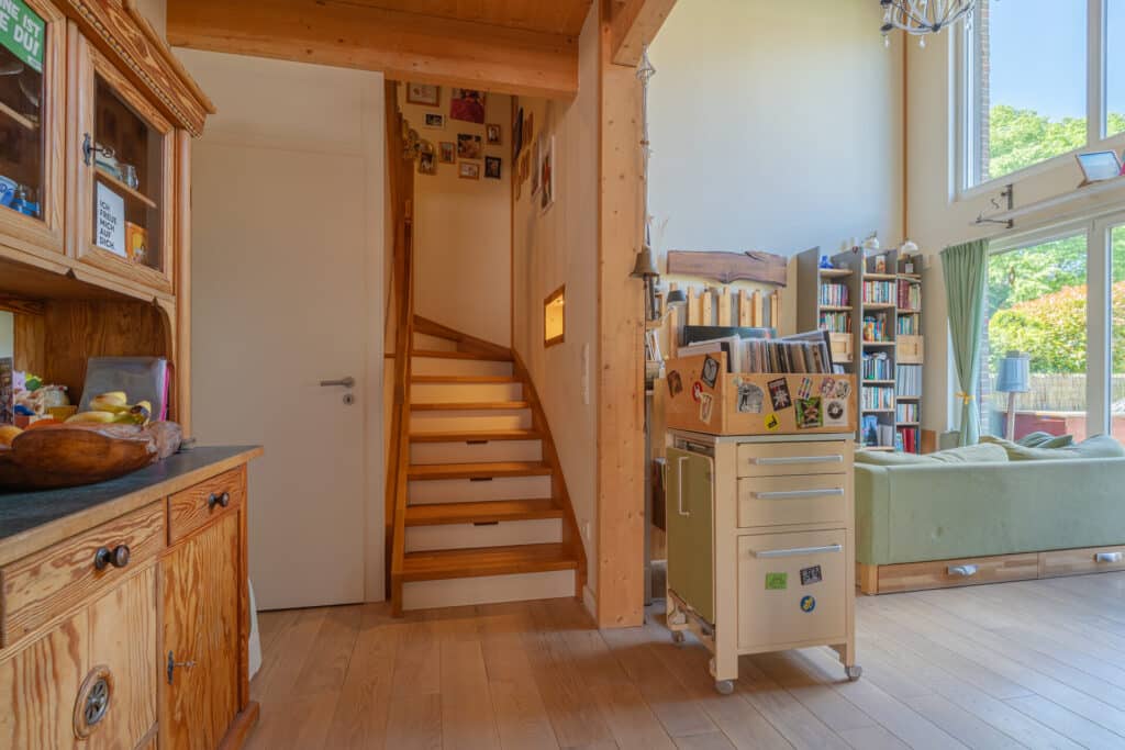 Ein behaglicher Wohnraum in einem nachhaltigen Fertighaus aus Holz mit warmen Holzböden, einer elegant geschwungenen Treppe ins Obergeschoss, viel Tageslicht durch großzügige Fenster, grünem Sofa, Bücherregalen und liebevollen Details für ein Zuhause zum Wohlfühlen.