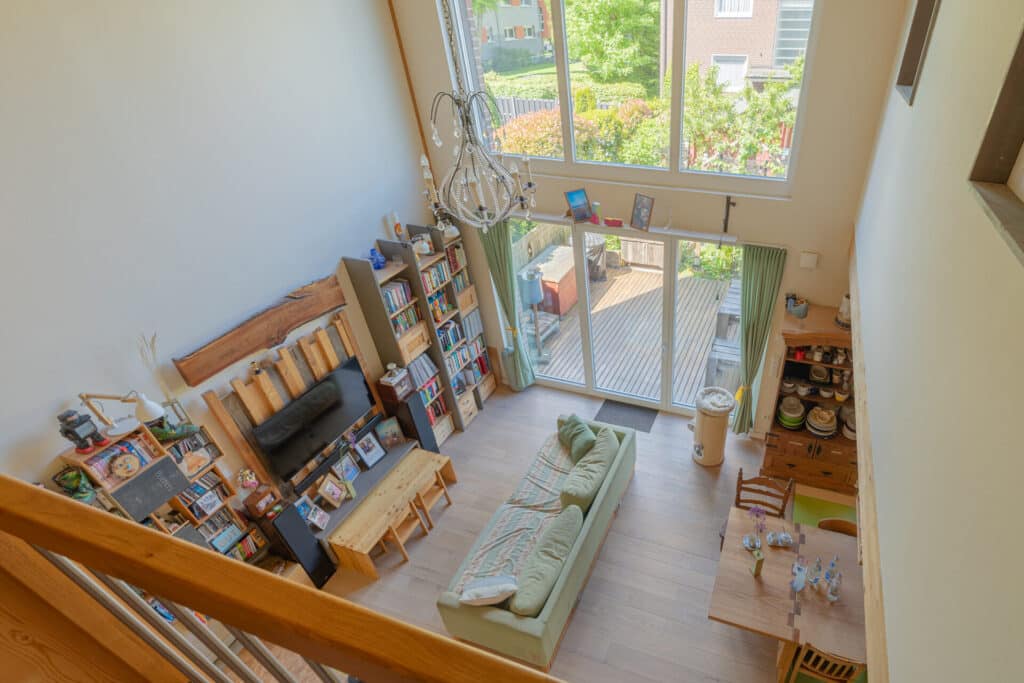 Einladendes, helles Wohnzimmer in einem modernen Holz-Fertighaus: Grünes Sofa und Holzmöbel schaffen mit großen Fenstern und natürlichem Licht eine behagliche Atmosphäre. Bücherregal, TV und Essbereich am Glaszugang zur Terrasse betonen den Wohnkomfort.