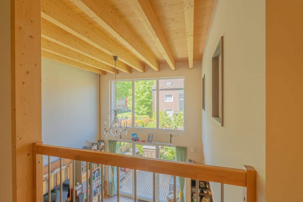 Blick aus einer oberen Galerie auf ein lichtdurchflutetes Wohnzimmer mit großen Fenstern, eleganter Holzdecke, Kronleuchter und Bücherregal. Natürliche Materialien und stilvolles Design schaffen Wohlfühlambiente; draußen grüne Natur und Nachbarhäuser.