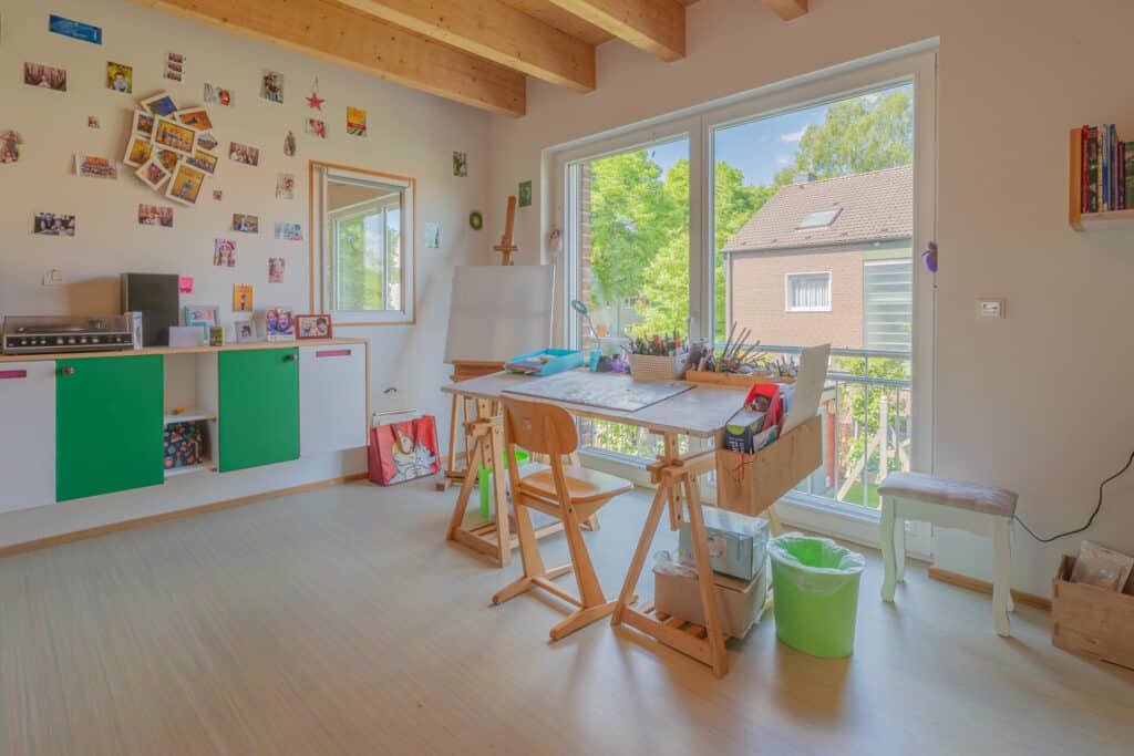 Ein lichtdurchflutetes Atelier mit großen Fenstern, sichtbaren Holzbalken und natürlichen Materialien schafft eine warme, behagliche Atmosphäre. Am Arbeitstisch stehen Stühle und Kunstmaterialien; bunte Werke zieren die Wände, draußen grünt es.