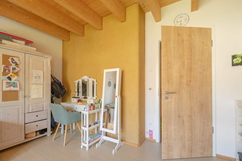 Ein behagliches Schlafzimmer in einem Fertighaus aus Holz, mit sichtbaren Holzbalken, warmer gelber Akzentwand, liebevoll dekoriertem Schrank mit Fotos, heller Schminkkommode samt Spiegel, hohem Standspiegel und Holztür mit Uhr - ein Raum zum Wohlfühlen.