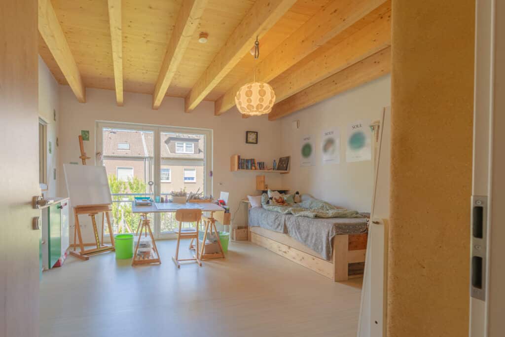 Helles, gemütliches Schlafzimmer mit sichtbaren Holzbalken in einem modernen Fertighaus aus Holz. Ein einzelnes Bett, Bücherregale und kreative Elemente stehen unter einem großen Fenster, das den behaglichen Raum mit natürlichem Licht durchflutet.