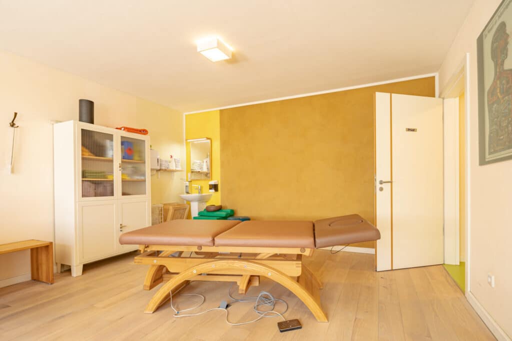 Ein heller Therapieraum mit leichter Holzliege, weißem Schrank und kleinem Waschbecken strahlt Wärme und Behaglichkeit aus. Helles Holzparkett und eine gelbe Akzentwand schaffen Wohlfühlatmosphäre, natürliche Materialien dominieren. Ein Telefon mit Kabel liegt am Boden.