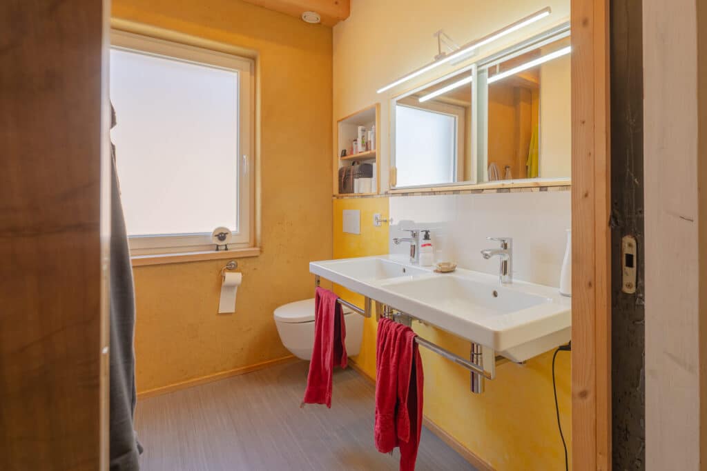 In einem Fertighaus aus Holz lädt dieses moderne Bad mit warmen gelben Wänden, großem satiniertem Fenster und Naturmaterialien zum Wohlfühlen ein. Zwei Waschbecken mit Spiegeln, rote Handtücher und dezente Ablagen sorgen für Komfort.