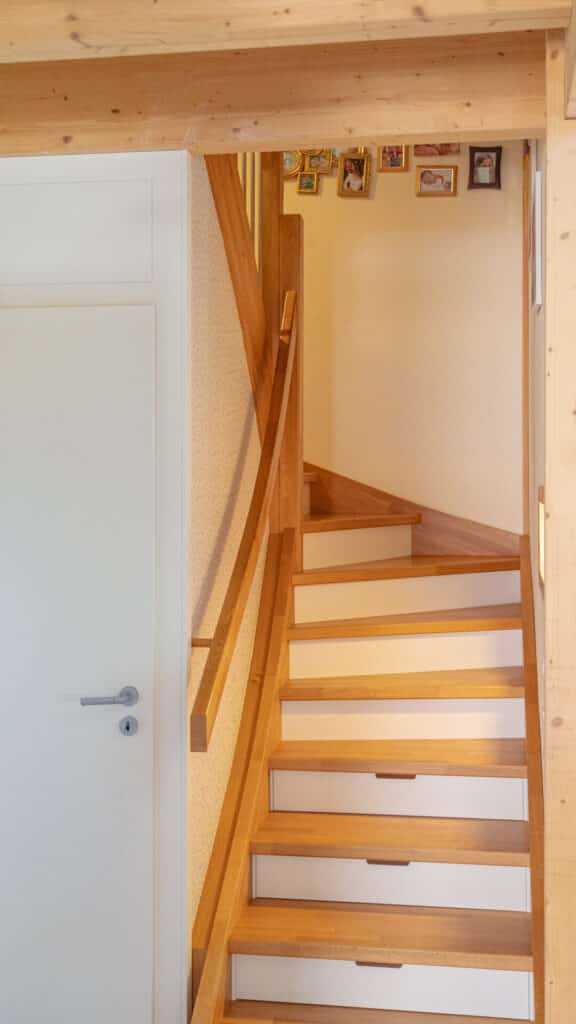 Eine elegante Holztreppe mit weißen Setzstufen windet sich nach oben und ist von massiven Holzbalken eingerahmt - ein Beispiel für nachhaltiges Wohnen, das Wärme und Geborgenheit ausstrahlt. Oben zieren gerahmte Fotos die Wand neben einer weißen Tür.