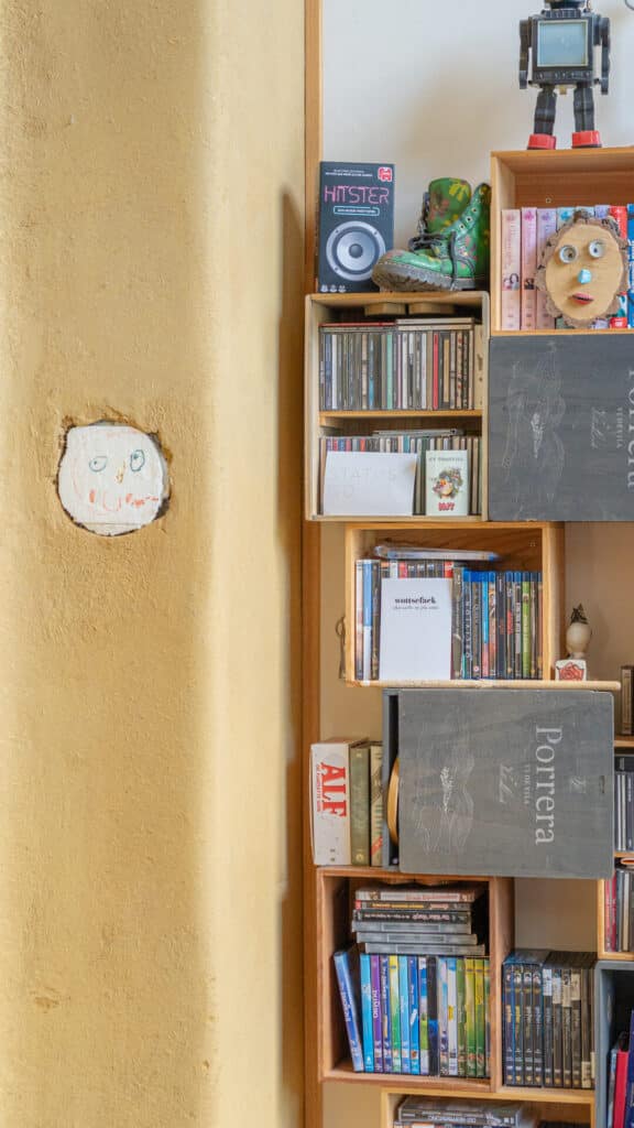 In unserem behaglichen Fertighaus aus Holz sorgen Bücherregale mit liebevoll ausgewählten Büchern, CDs und kreativen Deko-Elementen wie einer Roboterfigur für wohnliches Ambiente. Eine Wand ziert eine kindliche Zeichnung in sanften Farben.