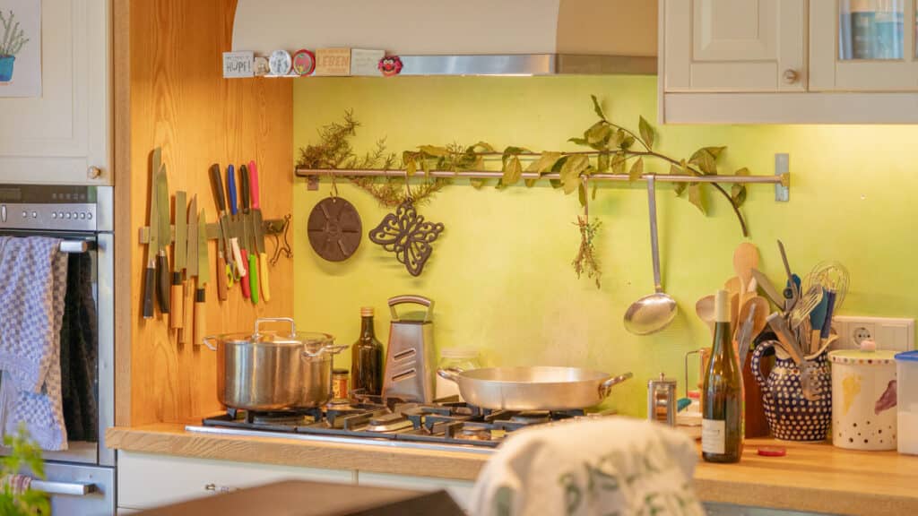 Eine behagliche Küche in einem modernen Fertighaus aus Holz vereint natürliche Materialien mit durchdachtem Design. Am Holzherd stehen Töpfe und Utensilien bereit, Messer hängen geordnet an der Wand, grüne Pflanzen und schöne Flaschen sorgen für wohnliches Ambiente.
