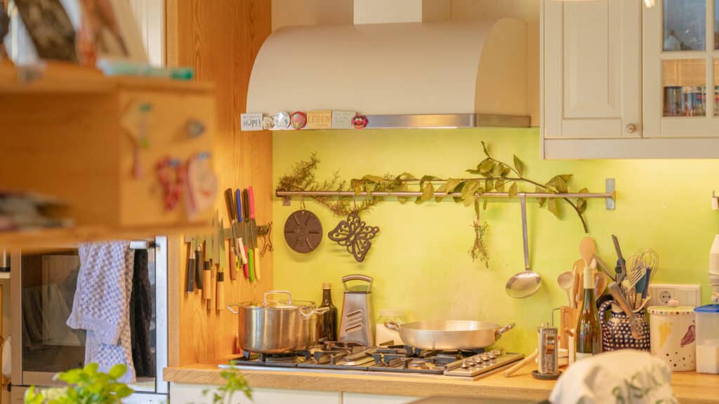 Einladende Küche mit grünen Wänden in einem nachhaltigen Fertighaus aus Holz. Natürliches Licht betont Regale, eine weiße Dunstabzugshaube und liebevoll arrangierte Details - ein perfektes Beispiel für wohngesundes, gemütliches Hausbau-Design.