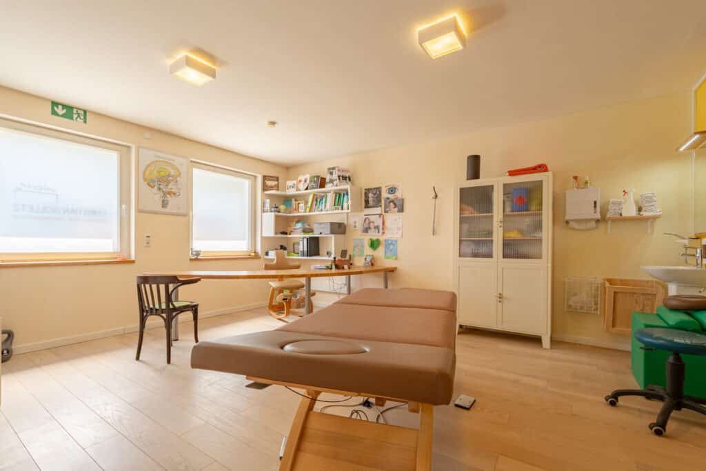Heller, einladender Behandlungsraum in einem nachhaltigen Fertighaus aus Holz: Viel Licht durch zwei Fenster, warme Gelbtöne an den Wänden, Holzboden, hochwertige Möbel - ein inspirierendes Beispiel für behaglichen Wohnkomfort und modernes Hausbau-Design.