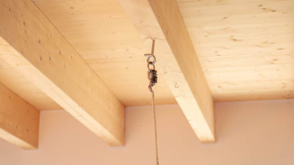Detailaufnahme einer Holzdecke mit sichtbaren Balken in einem behaglichen Fertighaus aus Holz. Ein hochwertiges Metallhaken- und Flaschenzugsystem mit herunterhängendem Seil unterstreicht den Mix aus Funktionalität, Komfort und natürlicher Wohnatmosphäre.