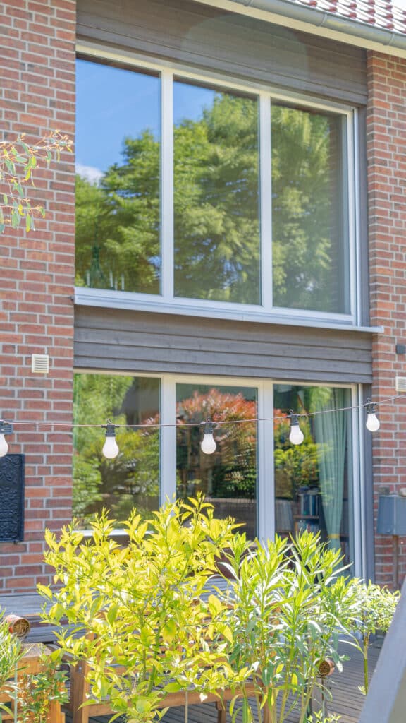 Ein nachhaltiges Fertighaus aus Holz mit großen Fenstern, gemütlicher Terrasse, stimmungsvollen Lichterketten und viel Grün im Vordergrund. Sonnenlicht und blauer Himmel verleihen dem Zuhause Wärme, Komfort und zeitlose Behaglichkeit.