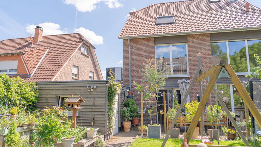 Ein sonniger Garten mit liebevoll gepflegten Pflanzen, einer Holzschaukel und einem Vogelhaus liegt zwischen zwei Backsteinhäusern mit roten Ziegeldächern - eine einladende Inspiration für nachhaltigen Hausbau im Grünen, der Behaglichkeit und modernes Design vereint.