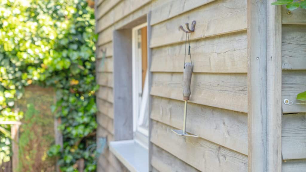 In einem lichtdurchfluteten Geräteschuppen aus Holz hängt ein Handrechen mit Holzgriff ordentlich an einem Haken an der Wand neben dem Fenster. Durch das Fenster schimmert grünes Laub, das für eine natürliche Wohlfühlatmosphäre sorgt.