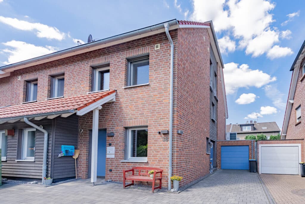 Ein modernes, teilmassives Haus mit großem grauen Fensterrahmen, rotem Ziegeldach und roter Bank am Eingang vermittelt Behaglichkeit. Zwei blaue Garagentore und eine gepflasterte Einfahrt ergänzen das harmonische Gesamtbild unter leicht bewölktem Himmel.