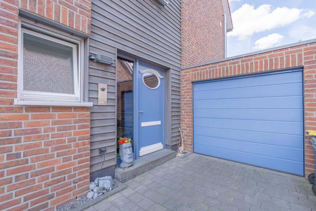 Eingang eines modernen Fertighauses aus Holz mit nachhaltiger Bauweise: Eine blaue Haustür mit rundem Fenster, passende Garagentür, Fassade aus Holz und Backstein sowie liebevoll arrangierte Blumentöpfe schaffen eine warme, einladende Atmosphäre.