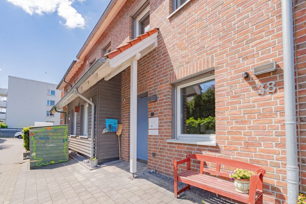 Ein einladendes Holz- und Backsteinhaus mit roter Bank, üppigem Pflanzkübel und überdachtem Eingang repräsentiert das nachhaltige Wohlfühlkonzept moderner Fertighäuser. Hausnummer 38 ist klar erkennbar, links befindet sich eine grüne Tonne.