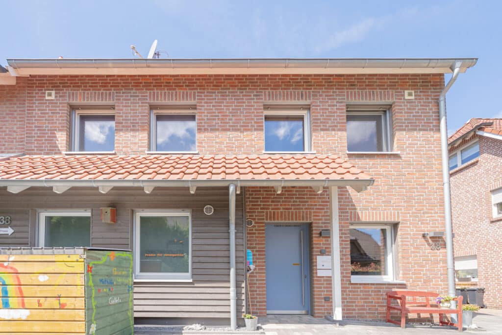 Ein modernes zweistöckiges Haus mit Ziegeldach, dessen natürliche Materialien und zeitgemäßes Design Behaglichkeit und Wärme vermitteln. Die blaue Haustür, ein passender Briefkasten sowie bunte Holzelemente und eine rote Bank betonen das einladende Ambiente.