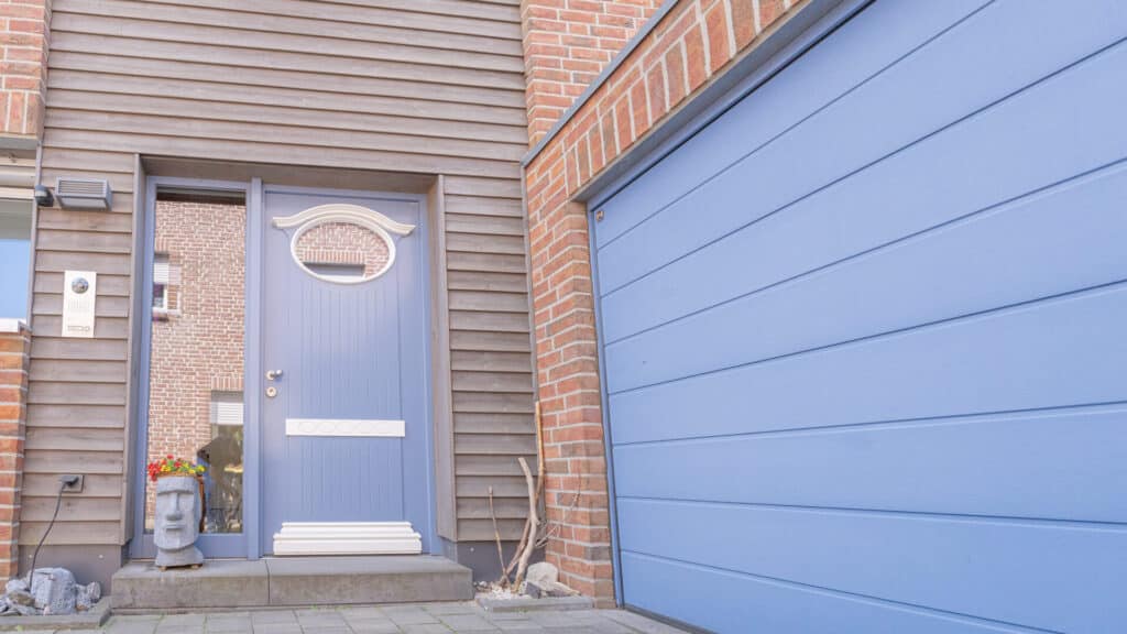 Ein hellblaues Eingangstor mit rundem Fenster und passendem Garagentor akzentuiert die Holz- und Klinkerfassade dieses Hauses. Blumen und eine Steinskulptur am Weg laden zum Wohlfühlen ein - ein Beispiel für nachhaltiges, behagliches Wohnen.