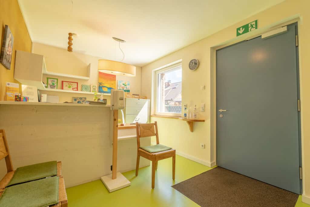 Ein heller, einladender Wartebereich mit grünem Boden, Holzmöbeln und warmen Akzenten wie Wandbildern und bunten Regalen. Die natürliche Holzoptik spiegelt das wohnliche Ambiente eines nachhaltigen Fertighauses wider.