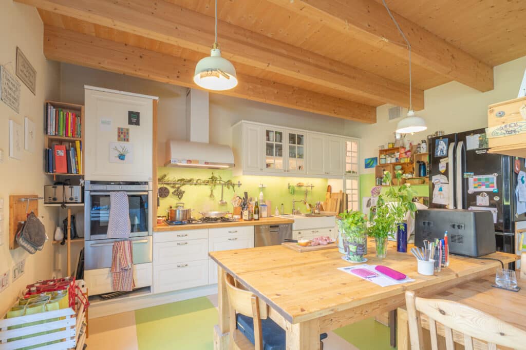 Ein helles, behagliches Holz-Fertighaus mit sichtbaren Holzbalken, weißen Küchenfronten und zentraler Insel. Pflanzen, Bücher und bunte Details verleihen dem Raum Wärme und laden dazu ein, sich im eigenen Traumhaus wohlzufühlen.