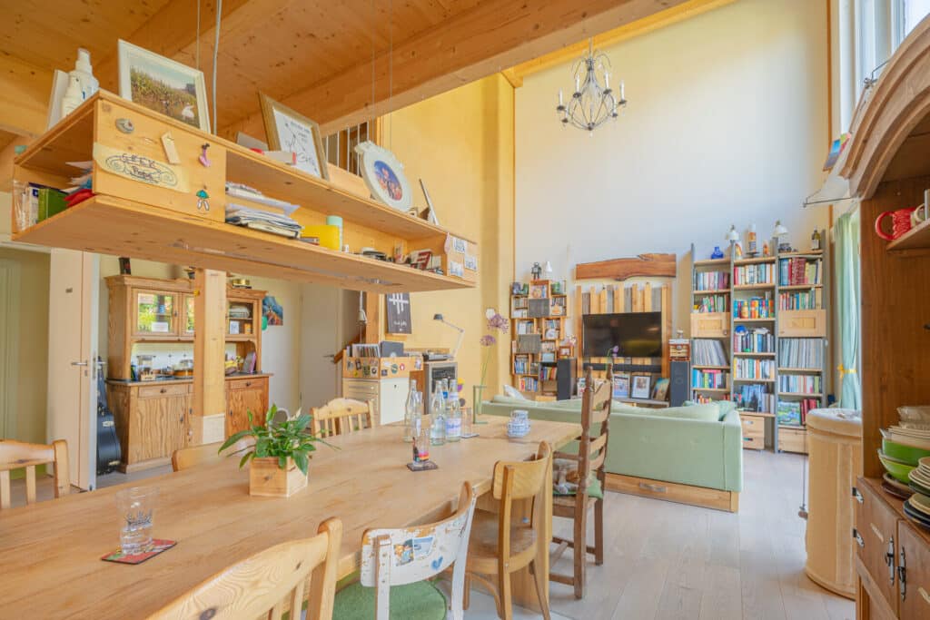 Sonnendurchfluteter Wohn- und Essbereich in einem Fertighaus aus Holz, mit natürlichen Holzdecken und -böden, großem Esstisch und verschiedenen Stühlen, grünen Sofas, Regalen, TV sowie liebevollen Details wie Pflanzen und Dekorationen - gemütlich und nachhaltig.