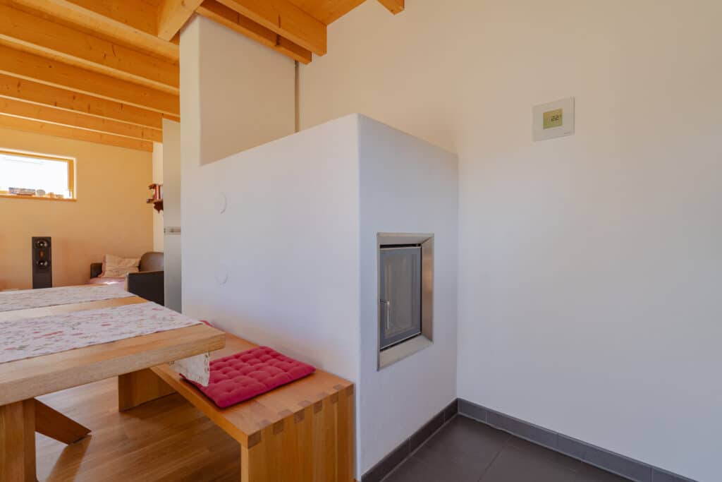 Ein lichtdurchfluteter, moderner Wohnraum mit Holz-Esstisch und Bänken mit roten Kissen, weißer Wand und sichtbaren Holzbalken - ein Beispiel für ein Fertighaus aus Holz, das Wärme, Behaglichkeit und nachhaltiges Design vereint.