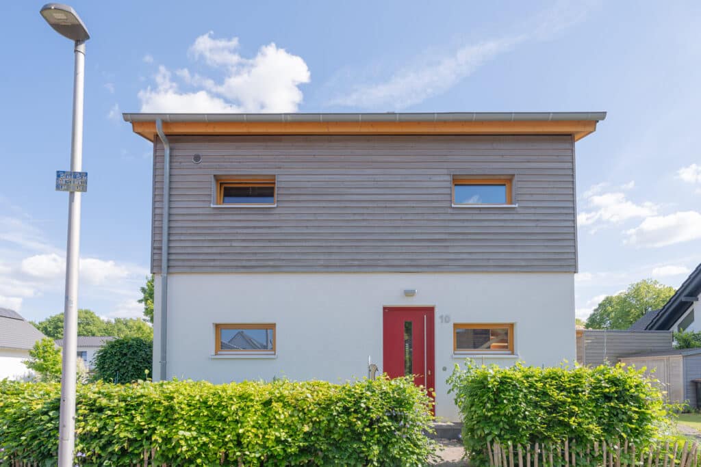 Unser zeitgemäßes, zweistöckiges Fertighaus aus Holz vereint natürliche Materialien mit modernem Design: Im Obergeschoss warme Holzverkleidung, darunter helle Wände, eine rote Eingangstür und quadratische Fenster - alles eingebettet in einen gepflegten Garten.
