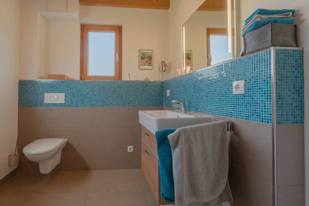 Ein modernes Bad in einem Hausbau-Projekt, gestaltet mit blauen Mosaikfliesen, wandhängendem WC und Waschbecken mit Holzunterschrank. Natürliches Licht durchflutet den Raum - Ausdruck nachhaltiger Bauweise, Komfort und zeitlosem Wohngenuss.