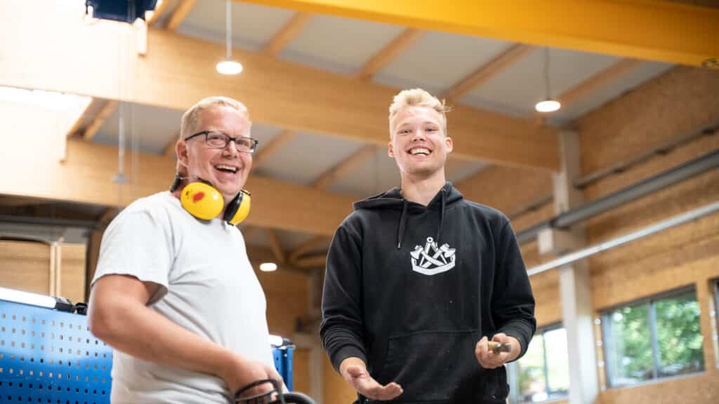 Zwei Männer stehen in einer lichtdurchfluteten, modernen Werkstatt und lächeln in die Kamera - ein Ort, an dem nachhaltige Fertighäuser aus Holz entstehen. Einer trägt Brille und Gehörschutz, der andere einen schwarzen Hoodie mit weißem Logo.