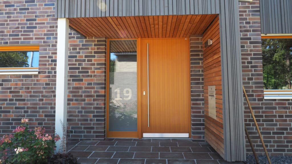 Der Eingang eines modernen Fertighauses aus Holz, mit großzügiger Holztür, hohem Seitenfenster samt Hausnummer 19, kombiniertem Mauerwerk und Holzverkleidung. Ein gefliester Weg und blühende Pflanzen schaffen eine einladende Wohlfühlatmosphäre.