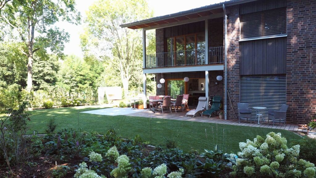 Ein modernes Haus mit Ziegel-Fassade, Balkon und Terrasse steht eingebettet in einem grünen Garten mit blühenden Blumen und Sträuchern. Der natürliche Holzbau fügt sich harmonisch in die umliegenden hohen Bäume ein und strahlt wohnliche Geborgenheit aus.