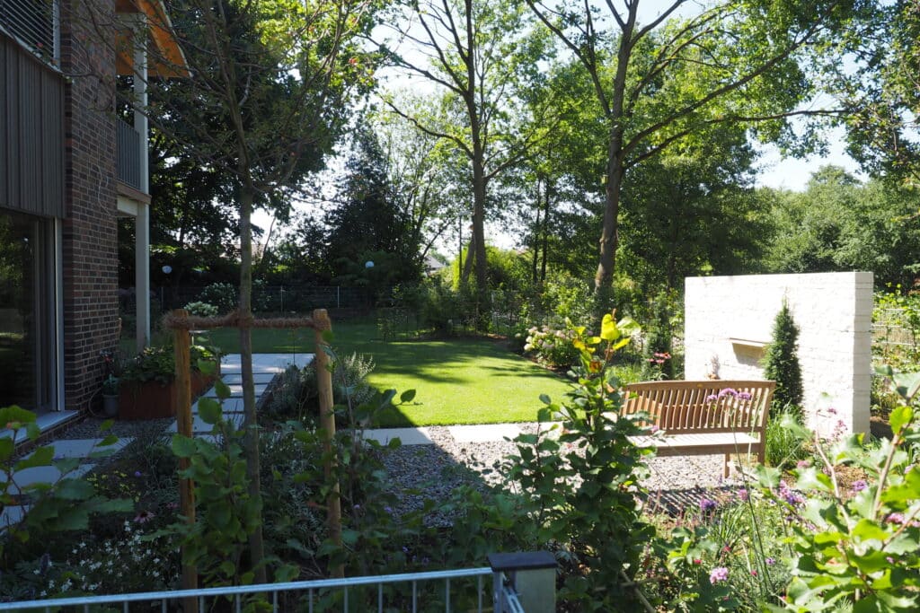 Ein sonniger Garten mit gepflegtem, grünem Rasen und farbenfrohen Blumen lädt zum Entspannen ein. Eine Holzbank vor einer weißen Steinmauer bietet einen gemütlichen Sitzplatz im Schatten hoher Bäume. Links zeigt sich ein Teil eines stilvollen Ziegelhauses.