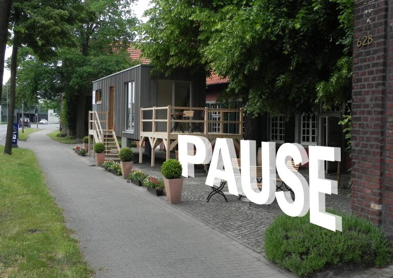 An einem begrünten Haus mit Tiny House ist das Wort PAUSE digital eingearbeitet.