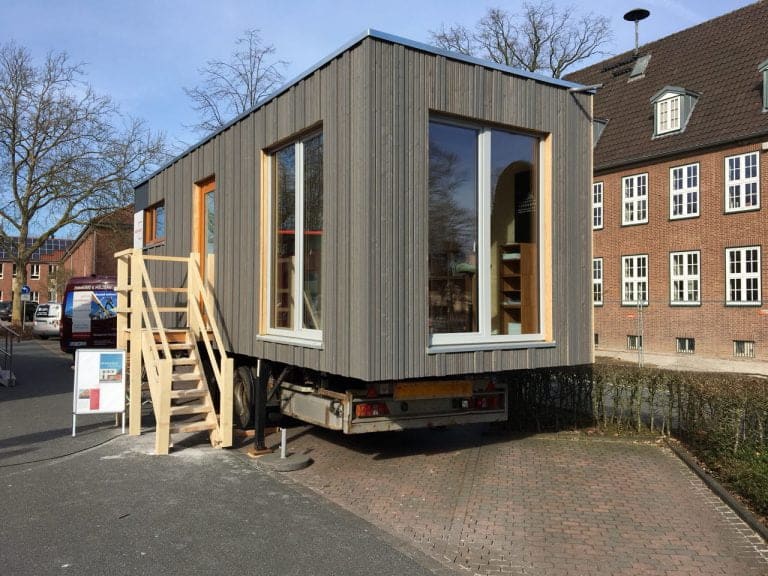 Ein Tiny House steht auf einen LKW-Anhänger vor einem Schulgebäude.