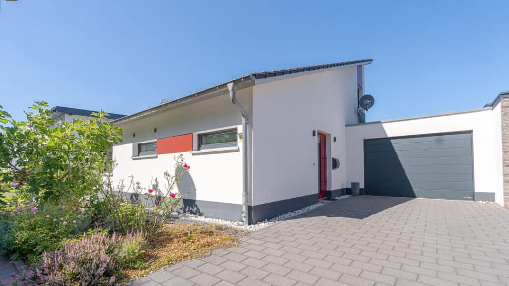 Ein modernes, eingeschossiges Holzfertighaus mit weißer Fassade, roten Akzenten und Flachdach lädt zum Wohlfühlen ein. Graues Garagentor, rote Haustür und eine kleine Blumenrabatte ergänzen das Gesamtbild eines hellen, behaglichen Zuhauses.