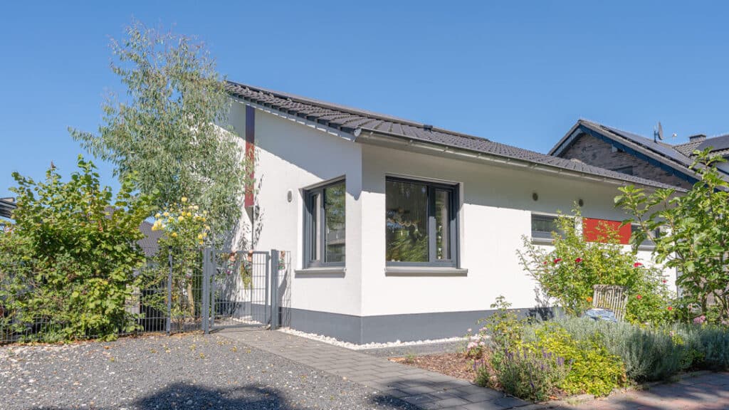 Modernes, eingeschossiges Holz-Fertighaus mit weißer Fassade, dunklem Dach und großen Fenstern, eingebettet in grüne Sträucher und Bäume unter blauem Himmel. Ein gepflasterter Weg führt zum einladenden Eingangsbereich.