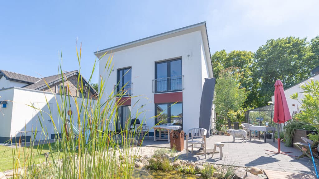 Nachhaltiges, modernes Zweifamilienhaus in Holzbauweise mit großen Fenstern und kleinem Balkon. Die sonnige Terrasse lädt mit gemütlichen Möbeln, einem roten Sonnenschirm und Blick auf den natürlichen Gartenteich zum Entspannen ein.