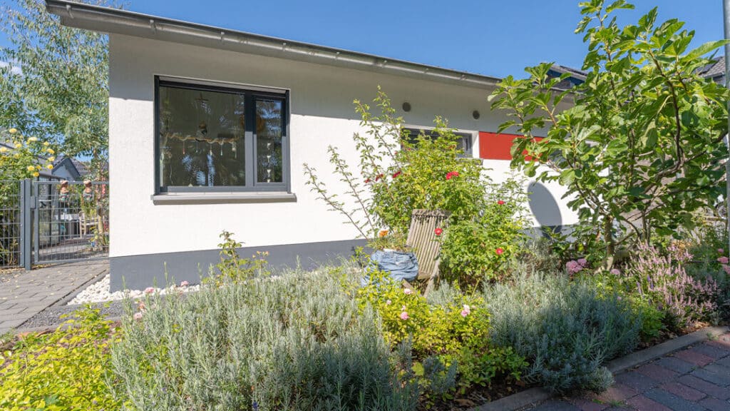 Unser modernes, eingeschossiges Holzfertighaus mit weißen Wänden und schwarzen Akzenten liegt eingebettet in einem liebevoll angelegten Garten mit Sträuchern, Blumen und kleinem Baum. Ein gepflasterter Weg führt durch dieses einladende Grün.