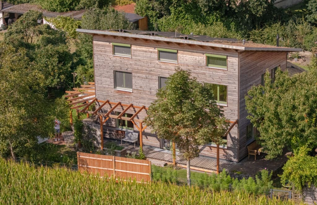 Ein modernes, zweistöckiges Holzhaus mit großen Fenstern und schrägem Dach bietet Geborgenheit inmitten von Bäumen und viel Grün. Die gemütliche Pergola mit Sitzbereich lädt im nachhaltigen, energieeffizienten Haus zum Wohlfühlen ein.